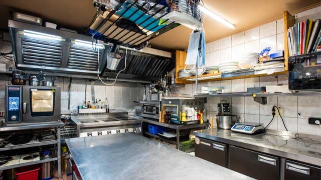 Interior of large kitchen of modern restaurant including big table for cooking, kitchenware, shelves, electric stove, oven and other equipment.